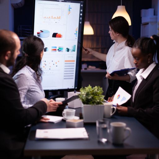Diverse multiethnic business teamwork overworking in office meeting room analyzing financial graphs late at night. Coworkers brainstorming project ideas solving company problem.
