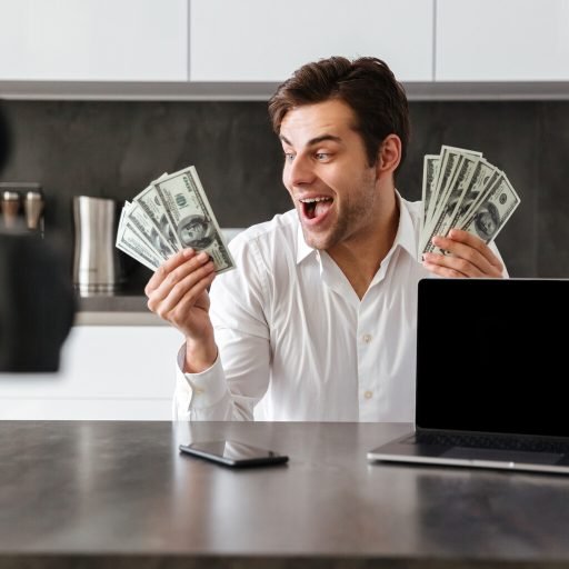Happy young man filming his video blog episode about new tech devices while sitting at the kitchen table with laptop and showing bunch of money banknotes